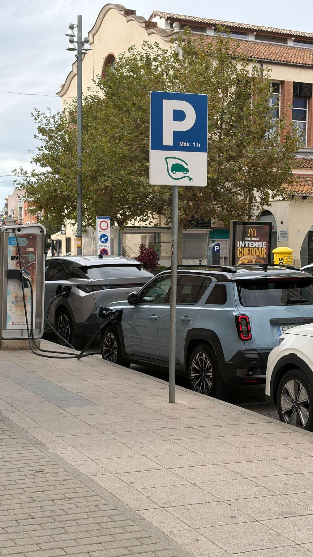 Renault 4 E-Tech azul claro enchufado a un punto de recarga público en la calle, junto a una señal de aparcamiento para vehículos eléctricos
